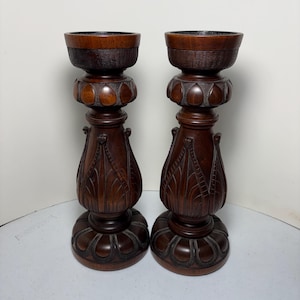 May include: Two dark brown wooden candle holders. Each has a round base, a carved stem with leaf-like details, and a bowl-shaped top. The wood grain is visible, and the design is ornate.