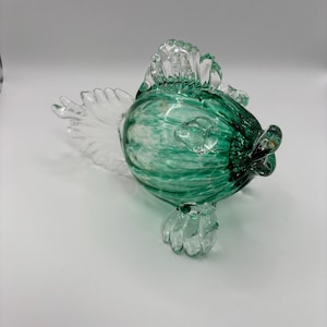 May include: A decorative glass fish sculpture. The fish features a rounded body with a mottled green pattern, clear fins, and a clear, textured tail. The sculpture is a unique piece of art.