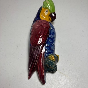 May include: A ceramic parrot wall hanging featuring a vibrant color palette of deep blue, maroon, yellow, and bright green. The bird is depicted perched on a small branch, with a dark brown beak, a yellow face, and textured plumage details.