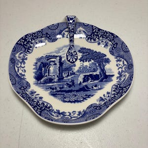 May include: A blue and white ceramic serving dish with a handle. The dish features a pastoral scene in the center and floral patterns around the rim. The handle is also decorated with blue floral designs.