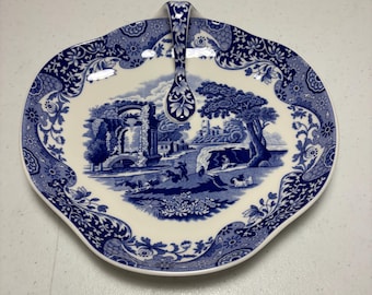 May include: A blue and white ceramic serving dish with a handle. The dish features a pastoral scene in the center and floral patterns around the rim. The handle is also decorated with blue floral designs.