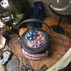 Puede incluir: Una bomba de ba&ntilde;o con forma de caldero negro con asa, llena de cristales de colores y un dado azul. El caldero tiene una etiqueta rosa que dice "Critical Cauldron bath bomb". El fondo incluye una calavera, una bolsa de terciopelo y monedas.