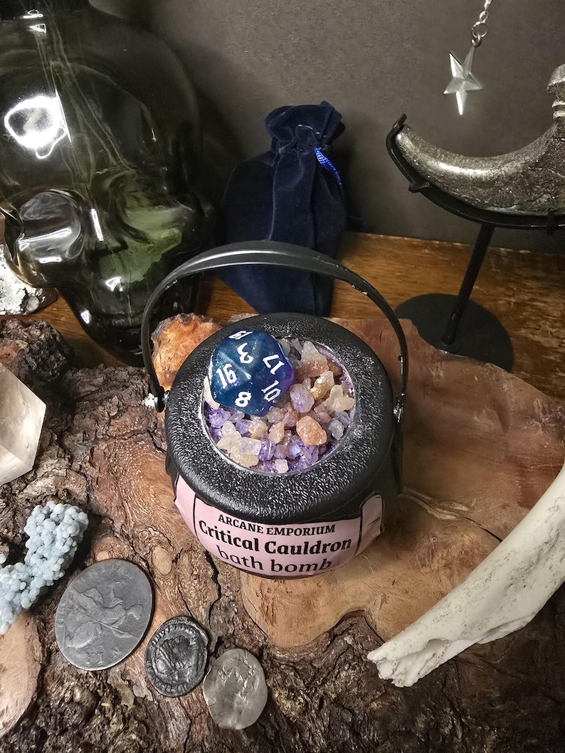 Puede incluir: Una bomba de ba&ntilde;o con forma de caldero negro con asa, llena de cristales de colores y un dado azul. El caldero tiene una etiqueta rosa que dice "Critical Cauldron bath bomb". El fondo incluye una calavera, una bolsa de terciopelo y monedas.