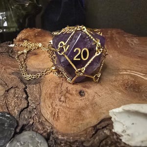 Könnte beinhalten: Eine lila, zwanzigseitige Würfel-Anhänger-Halskette mit einer goldfarbenen Kette. Der Würfel besteht aus einem durchscheinenden lila Material und hat die Zahl 20 in Gold. Der Würfel ist in einem goldfarbenen Metallkäfig eingeschlossen. Die Halskette liegt auf einem Holzstück.
