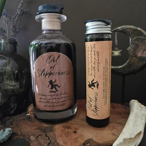 May include: Two glass bottles labeled "Oil of Slipperiness" with dark liquid inside. The larger bottle has a dark blue stopper and a brown label with text and a falling figure. The smaller bottle has a silver cap and a similar label.