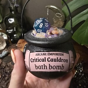 Puede incluir: Una bomba de ba&ntilde;o negra con forma de caldero con asa, llena de cristales y un dado azul y blanco. La etiqueta dice "Arcane Emporium Critical Cauldron bath bomb."
