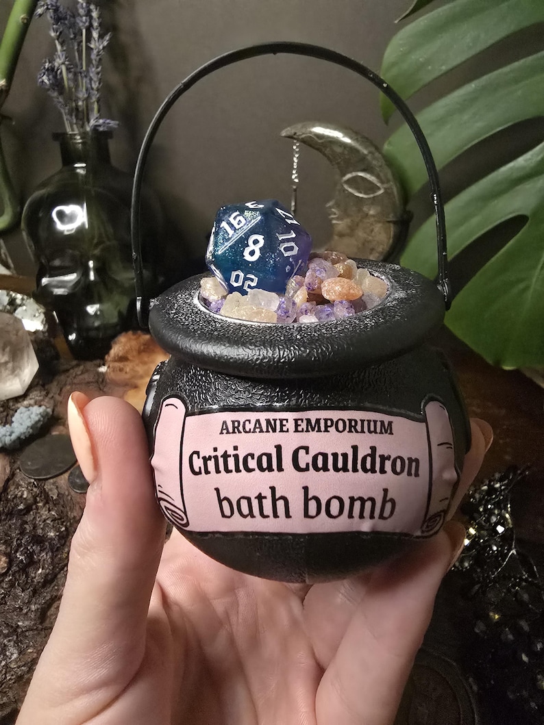 Puede incluir: Una bomba de ba&ntilde;o negra con forma de caldero con asa, llena de cristales y un dado azul y blanco. La etiqueta dice "Arcane Emporium Critical Cauldron bath bomb."