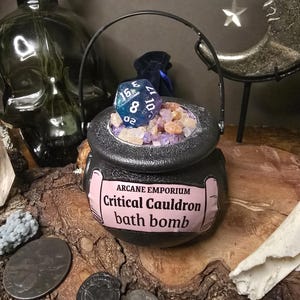 Puede incluir: Una bomba de ba&ntilde;o de caldero negro etiquetada como "Arcane Emporium Critical Cauldron bath bomb", llena de cristales coloridos y un dado de 20 caras azul y blanco. El caldero tiene un asa negra y est&aacute; sobre una superficie de madera.