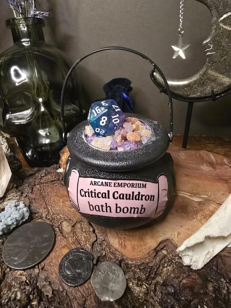 Puede incluir: Una bomba de ba&ntilde;o de caldero negro etiquetada como "Arcane Emporium Critical Cauldron bath bomb", llena de cristales coloridos y un dado de 20 caras azul y blanco. El caldero tiene un asa negra y est&aacute; sobre una superficie de madera.