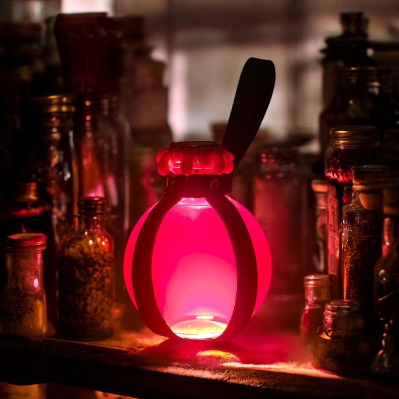 K&ouml;nnte beinhalten: Eine rote Glasflasche mit einem schwarzen Ledergriff und einem roten Metallk&auml;fig. Die Flasche ist mit einer leuchtend roten Fl&uuml;ssigkeit gef&uuml;llt und von anderen Flaschen umgeben.
