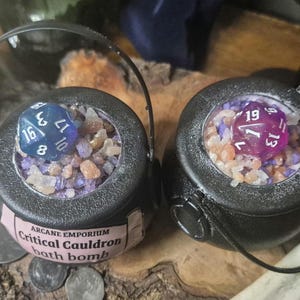 Puede incluir: Dos bombas de ba&ntilde;o negras en forma de caldero, cada una llena de cristales de colores y un dado grande. Un dado es azul, el otro es morado. Las bombas de ba&ntilde;o tienen una etiqueta que dice "Arcane Emporium Critical Cauldron bath bomb."