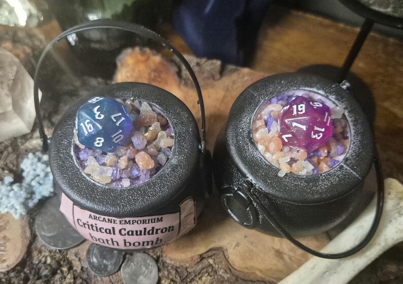 Puede incluir: Dos bombas de ba&ntilde;o negras en forma de caldero, cada una llena de cristales de colores y un dado grande. Un dado es azul, el otro es morado. Las bombas de ba&ntilde;o tienen una etiqueta que dice "Arcane Emporium Critical Cauldron bath bomb."