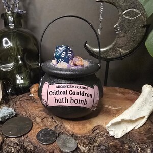 Puede incluir: Una bomba de ba&ntilde;o de caldero negro con una etiqueta rosa que dice "Critical Cauldron bath bomb". El caldero est&aacute; lleno de cristales y un dado azul y turquesa. La imagen tambi&eacute;n incluye un jarr&oacute;n con forma de calavera y una decoraci&oacute;n de media luna.