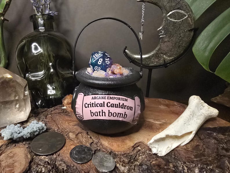 Puede incluir: Una bomba de ba&ntilde;o de caldero negro con una etiqueta rosa que dice "Critical Cauldron bath bomb". El caldero est&aacute; lleno de cristales y un dado azul y turquesa. La imagen tambi&eacute;n incluye un jarr&oacute;n con forma de calavera y una decoraci&oacute;n de media luna.