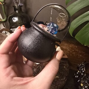 Puede incluir: Un peque&ntilde;o caldero negro con asa, lleno de cristales y un dado azul. Un dije de media luna cuelga cerca. El caldero se sostiene en una mano, con otros elementos decorativos en el fondo, incluyendo un jarr&oacute;n con lavanda.