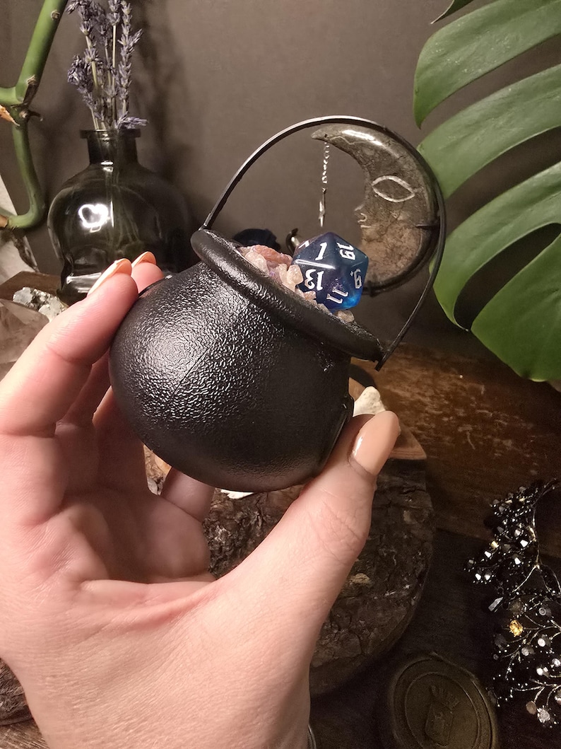 Puede incluir: Un peque&ntilde;o caldero negro con asa, lleno de cristales y un dado azul. Un dije de media luna cuelga cerca. El caldero se sostiene en una mano, con otros elementos decorativos en el fondo, incluyendo un jarr&oacute;n con lavanda.