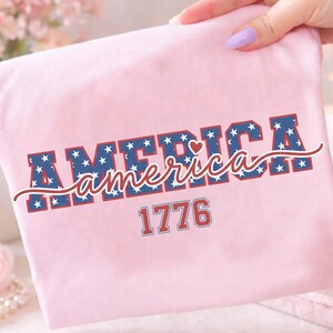 May include: A light pink t-shirt with the word "AMERICA" in a bold, blue font filled with white stars. The word "America" is written in a cursive font in red. The number "1776" is printed below in a red font.