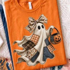 May include: An orange t-shirt featuring a graphic of a ghost made of various fabric patches, including plaid and floral patterns. The ghost wears a large matching fabric bow on its head and carries a small pumpkin-shaped trick-or-treat basket.