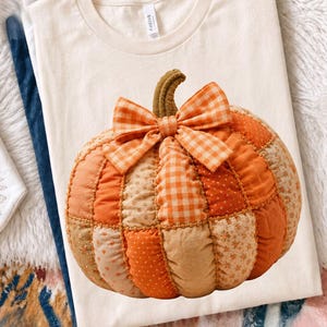 以下が含まれることがあります： クリーム色のTシャツで、中央に詰め物をしたキルティング加工の大きなカボチャのグラフィックが描かれています。カボチャはオレンジやベージュの布地をパッチワーク状に組み合わせたもので、花柄やドット柄があしらわれ、ギンガムチェックのリボンが添えられています。