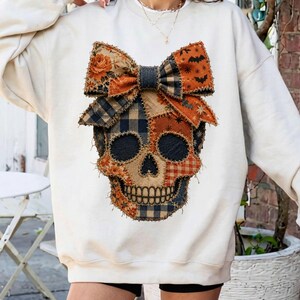 Peut inclure: Un sweat-shirt à manches longues de couleur crème arborant un grand motif central représentant un crâne orné d'un nœud imposant. Le crâne et le nœud sont composés de divers motifs en patchwork dans des tons orange, noir et beige.