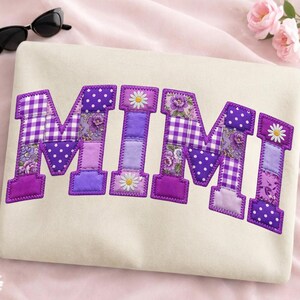May include: Cream-colored sweatshirt with the name "MIMI" appliqued in patchwork style. The letters are purple with floral, polka dot, and gingham patterns. Each letter is outlined with a purple zigzag stitch. Sunglasses are in the upper left corner.