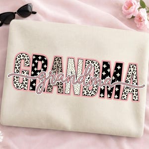 May include: A cream-colored sweatshirt with the word "GRANDMA" in a bold, decorative font. The letters have a leopard print and black cross design, outlined in pink. The word "grandma" is written in a cursive font over the word "GRANDMA".