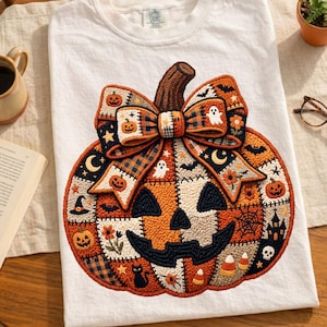 以下が含まれることがあります： 白いTシャツに刺繍されたカボチャのデザイン。カボチャはオレンジ色で、黒いジャック・オ・ランタンの顔と茶色の茎があります。チェック柄のリボンと、お化けやコウモリなど、ハロウィーンをテーマにしたさまざまなパッチがデザインを完成させています。