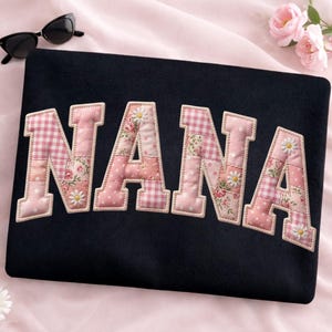Könnte beinhalten: Eine marineblaue Laptoptasche mit dem Namen "NANA" aus rosa Patchworkstoff. Die Buchstaben haben Blumen-, Vichy- und Tupfenmuster mit Gänseblümchen-Akzenten. Eine Sonnenbrille liegt oben links.