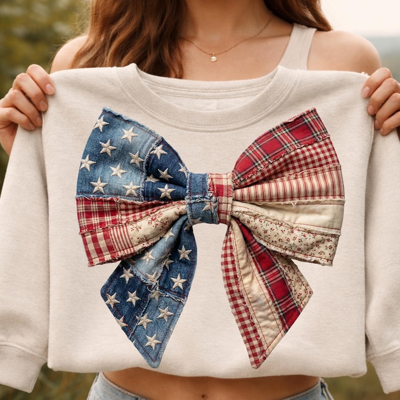 May include: A cream-coloured sweatshirt with a large bow design. The bow is a patchwork of denim with white stars, red plaid, and floral fabric. The design evokes a patriotic theme.