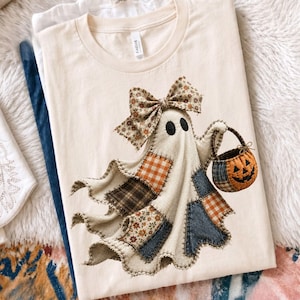Op de afbeelding: Een cr&egrave;mekleurig t-shirt met een afbeelding van een geest gemaakt van diverse stoffen lapjes, waaronder oranje gingham, bruine ruitjes, denim en bloemenprints. De geest draagt een grote bijpassende strik en draagt een kleine pompoenemmer.