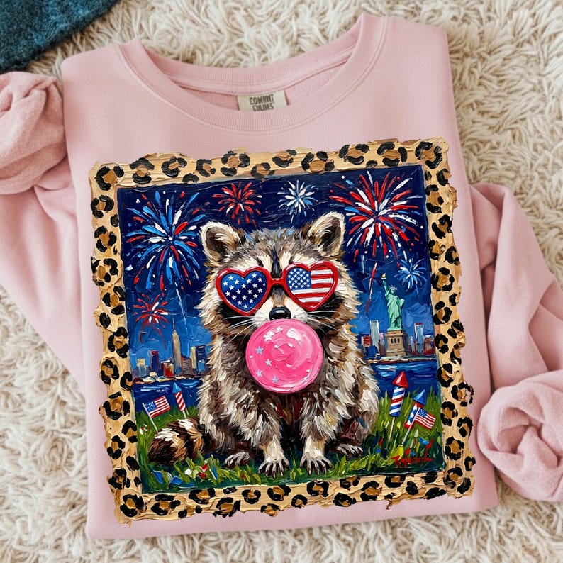 May include: Pink crewneck sweatshirt featuring a raccoon wearing heart-shaped sunglasses with an American flag design, blowing a pink bubble. The graphic includes fireworks, the Statue of Liberty, and a cityscape. The image is framed with a leopard print border.