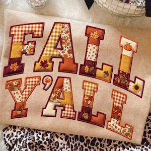 May include: A beige sweatshirt featuring the text FALL Y'ALL in large, block-style letters. Each letter is filled with a patchwork of autumn-themed fabric patterns, including orange and white gingham, sunflowers, pumpkins, leaves, and acorns.