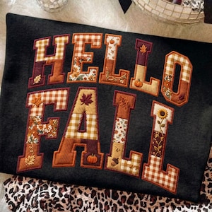 Peut inclure: Un sweat-shirt noir avec le texte HELLO FALL en grandes lettres majuscules. Chaque lettre est remplie d'un patchwork de tissus aux motifs automnaux, incluant du vichy orange et blanc, des fleurs, des citrouilles, des feuilles et des tournesols.