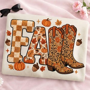 K&ouml;nnte beinhalten: Cremefarbenes Sweatshirt mit dem gestickten Wort "FALL". Das "F" hat ein braun-beiges Karomuster, das "A" ist mit Herbstbl&auml;ttern und K&uuml;rbissen gef&uuml;llt, und das "LL" sind ein Paar Cowboy-Stiefel mit floralen Designs. Eicheln und K&uuml;rbisse sind verstreut.