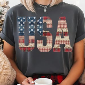 May include: Dark grey t-shirt with the word "USA" in a patchwork design. The letters are made from denim, red plaid, and floral patterns, resembling the American flag. The shirt has rolled-up sleeves.