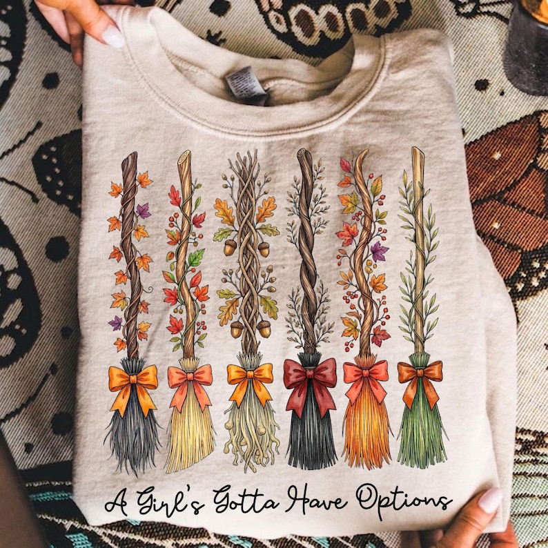 May include: Beige sweatshirt featuring a design of six decorative witch brooms with colorful autumn leaves and bows. The brooms have different colored bristles and bows. The text "A Girl's Gotta Have Options" is printed at the bottom.