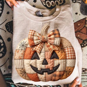 May include: A beige sweatshirt with a large, patchwork pumpkin applique. The pumpkin features a smiling jack-o'-lantern face, a bow, and spiderweb details. The pumpkin is made of various fabrics in orange, brown, and cream colors.