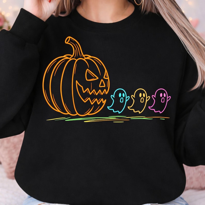 Op de afbeelding: Zwart sweatshirt met een neonoranje pompoenontwerp en drie kleurrijke spookfiguren. Het ontwerp bevat een neon groene, gele en roze lijn aan de onderkant, waardoor een Halloween-thema ontstaat.
