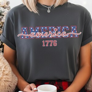 May include: Dark gray t-shirt with the word "America" in blue letters filled with white stars, and a red heart. Below the word "America" is the number "1776" in red. The shirt has rolled-up sleeves.