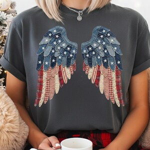 May include: Dark grey t-shirt with a design of denim and plaid wings with white stars. The wings are made from blue denim and red and white plaid fabric. The shirt has short sleeves and a relaxed fit.
