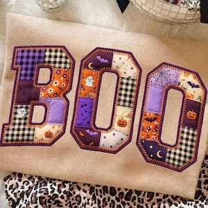 May include: Three fabric letters spelling BOO, each featuring a patchwork design with purple, orange, black, and white patterns. The letters include Halloween-themed motifs such as ghosts, pumpkins, bats, stars, and spiderwebs, all outlined with a decorative orange stitched border.
