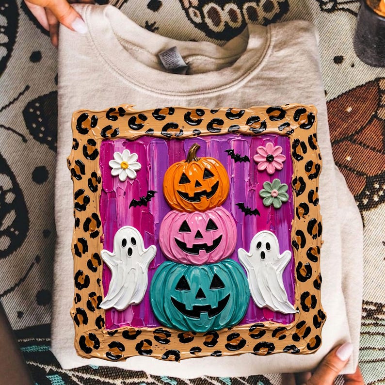 Puede incluir: Una sudadera beige de cuello redondo con un gr&aacute;fico cuadrado con borde de estampado de leopardo. El centro muestra una pila de tres calabazas de Halloween en naranja, rosa y turquesa, flanqueadas por dos fantasmas blancos y flores.