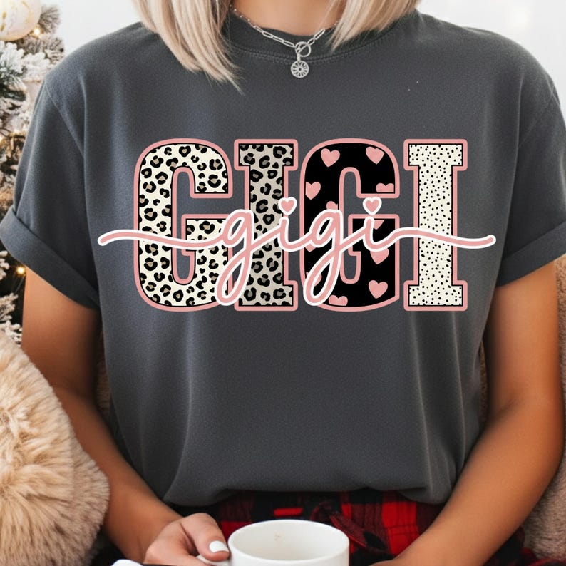 May include: Dark gray t-shirt featuring the word "GIGI" in a bold, decorative font. The letters have leopard print, heart, and polka dot patterns with a pink outline. The word "gigi" is written in a cursive font across the main text.