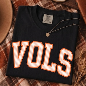 May include: A black short-sleeved t-shirt featuring the word VOLS in large, block-style letters. The letters are white with thin vertical orange stripes and a solid orange outline. The shirt has a Comfort Colors brand label at the inner neckline.