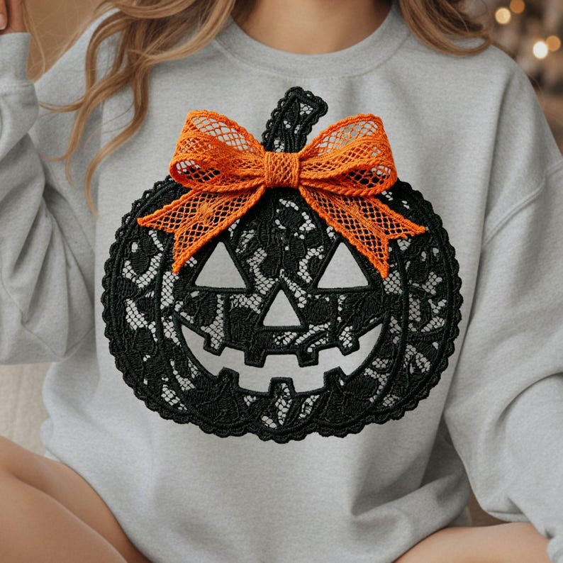 Może przedstawiać: Jasnoszara bluza z okrągłym dekoltem, ozdobiona z przodu dużą grafiką dyni wykonaną z czarnej koronki. Dynia ma twarz jack-o'-lantern z tr&oacute;jkątnymi oczami i nosem oraz jest ozdobiona jaskrawopomarańczową koronkową kokardą przy ogonku.
