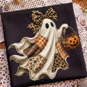 May include: A fabric ghost ornament featuring a cream-colored body with black stitched edges, adorned with a leopard print bow. The ghost holds a small orange pumpkin-shaped basket with a black jack-o'-lantern face and various fabric patches.