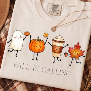 May include: A cream-colored t-shirt featuring a graphic of a ghost, a pumpkin, a coffee cup, and a maple leaf with faces. The text "FALL IS CALLING" is printed below the characters. A gold-toned necklace rests on the neckline of the shirt.