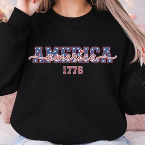 May include: Black sweatshirt with the word "AMERICA" in a red, white, and blue star-patterned font. The word "America" is in a cursive font. Below the word "America" is the number "1776" in a red font.