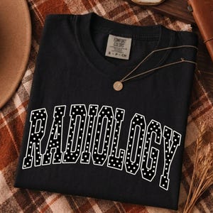 May include: A black short-sleeved t-shirt featuring the word RADIOLOGY printed in a large, white, varsity-style font with a polka dot pattern. The shirt has a crew neckline and a Comfort Colors brand label on the inside collar showing size small.