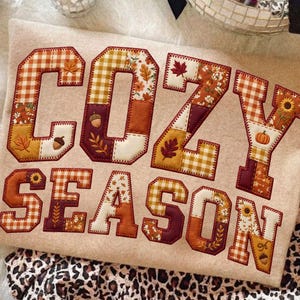 May include: A beige sweatshirt featuring the text COZY SEASON in large, block-style letters. Each letter is filled with a patchwork pattern of orange, yellow, and brown autumn-themed fabrics, including gingham, sunflowers, leaves, and acorns.